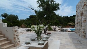 An outdoor dining area with a stone table set for six, surrounded by stone walls and a pool in the background. - Architecturally Designed Sea View Villa in Carovigno, Nr Ostuni with Heated Pool (carovigno)