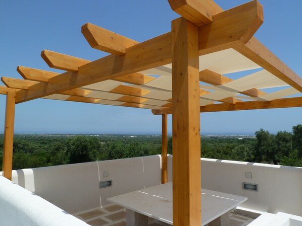 A wooden pergola structure with a table and seating area, overlooking a green landscape and distant horizon.