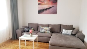 A modern living room with a sectional sofa, a small white table with a vase of flowers, and a large painting of a sunset over water on the wall.