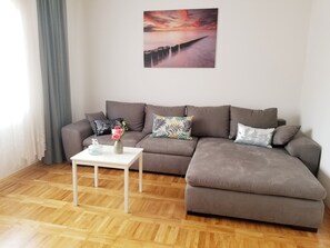 Una sala de estar moderna con un sofá seccional, una mesita blanca con un jarrón de flores y un cuadro grande de un atardecer sobre el agua en la pared.