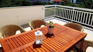 A wooden table with wicker chairs on a balcony with a white railing, overlooking a coastal area with houses and mountains in the background.