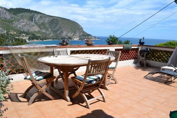 Une terrasse dotée d’une table ronde et de chaises, offrant une vue sur un paysage côtier avec des falaises et la mer.