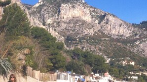 Una zona costera con una montaña rocosa al fondo, una playa con algunos edificios y una persona de pie sobre la arena.
