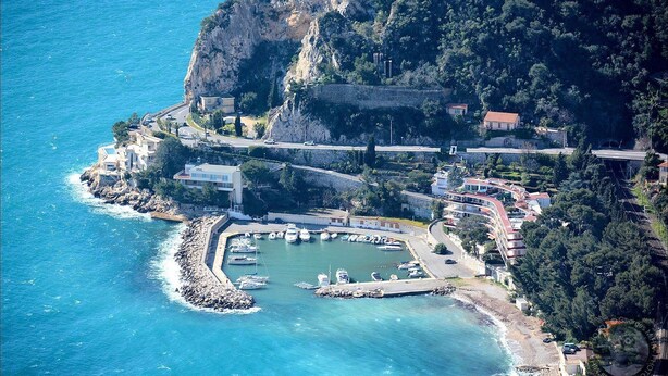 Vista aérea de un pueblo costero con un puerto deportivo, edificios y un litoral rocoso.