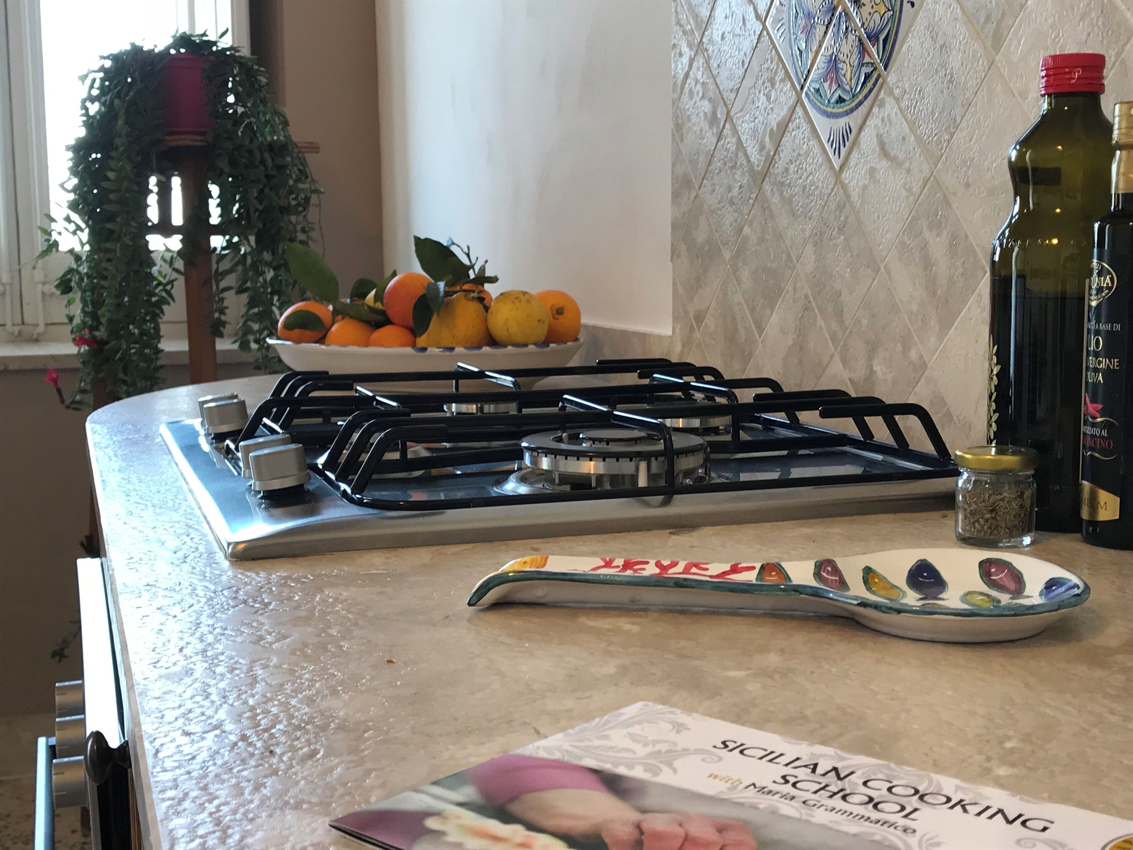 A kitchen counter with a gas stove, a bowl of fruit, a cookbook, and a bottle of olive oil.