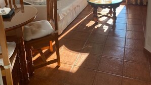 Un espacio de sala acogedor con sofá, una mesa de comedor y sillas. Hay una lámpara de techo y una lámpara de pie. El cuarto tiene piso de baldosas y una puerta corrediza de vidrio que da a un área exterior.