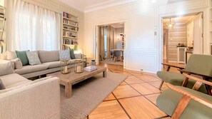 Ein Wohnzimmer mit einer Couch, einem Couchtisch und Sesseln, einem Bücherregal und einem Essbereich im Hintergrund.