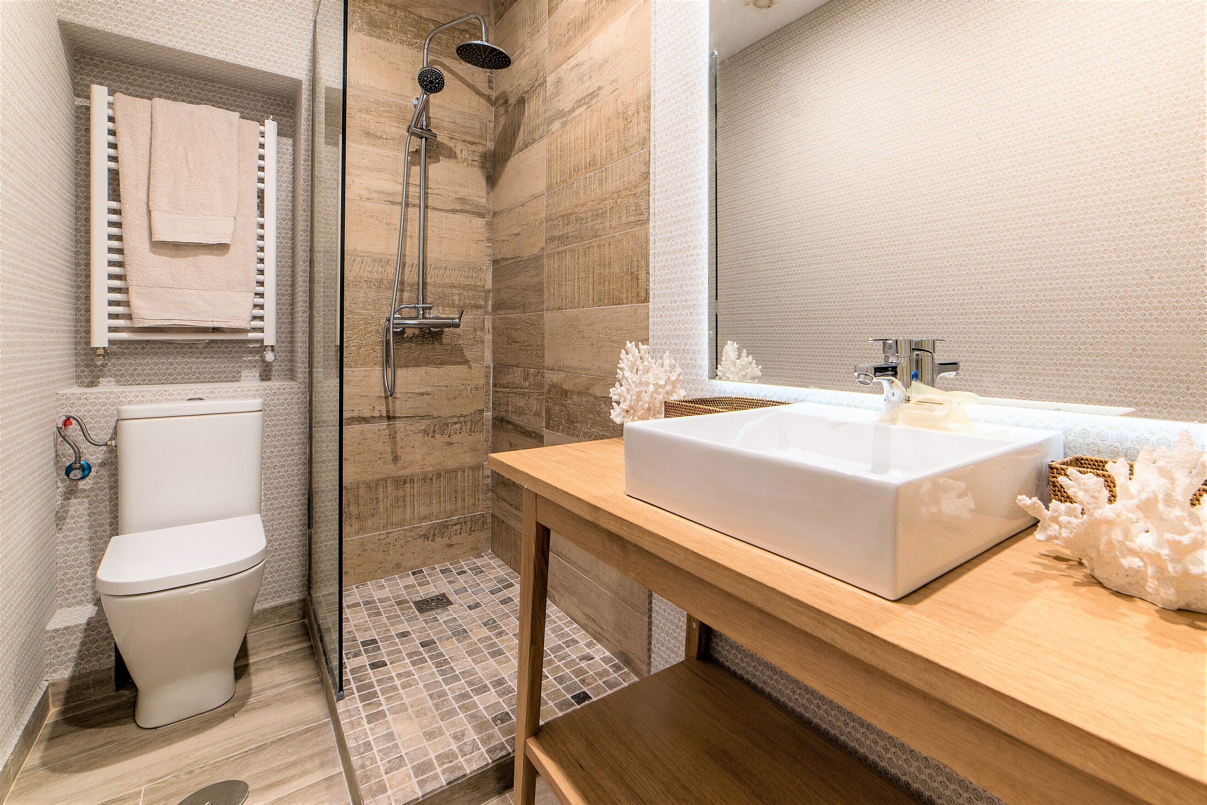 Un baño moderno con inodoro blanco, ducha con cerramiento de vidrio y tocador de madera con un lavamanos grande y blanco.