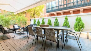 Un patio al aire libre con una mesa y sillas, rodeado de follaje y la fachada de un edificio.