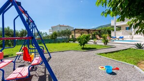 A playground with swings and a slide, a pool, and a building with a balcony. - Luxury Villa With Pool, Gym, Bicycles And Beautiful Sea View (Kaštel Sucurac)