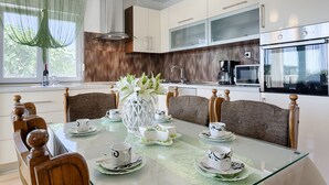 A modern kitchen with white cabinets, a microwave, and a coffee maker. There is a dining table with chairs and a vase of flowers. - Luxury Villa With Pool, Gym, Bicycles And Beautiful Sea View (Kaštel Sucurac)