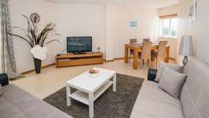 Ein modernes Wohnzimmer mit einer Couch, einem Couchtisch, einem Fernseher, einem Esstisch mit Stühlen und einem Fenster.
