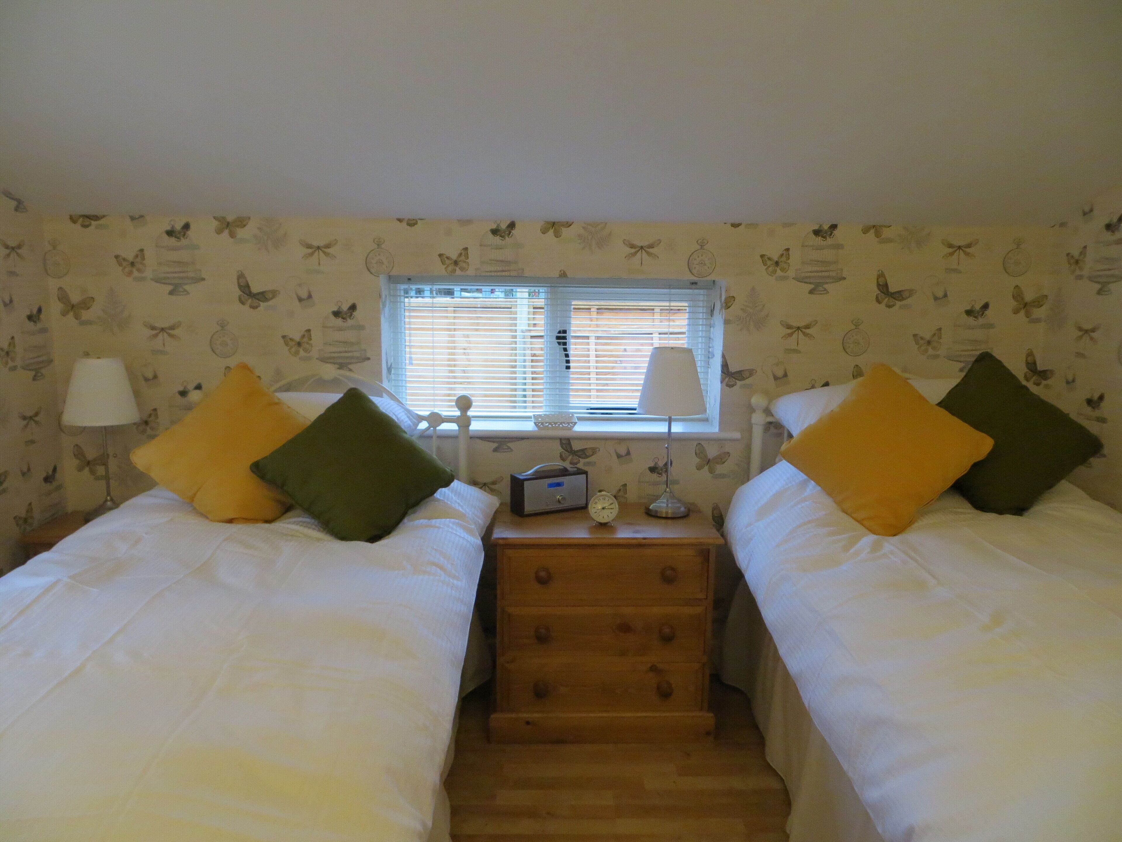 A bedroom with two beds, each with a pillow, a nightstand with a lamp, a clock, and a window with blinds.