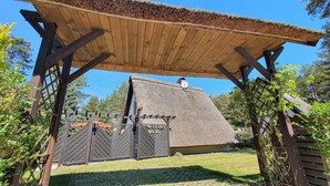 Une pergola en bois avec un toit de chaume, une zone délimitée par une clôture et un bâtiment doté d’un toit en pente.