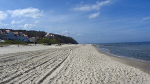 Une plage de sable avec des empreintes de pas menant à l’eau, un ciel dégagé et des bâtiments en arrière-plan.