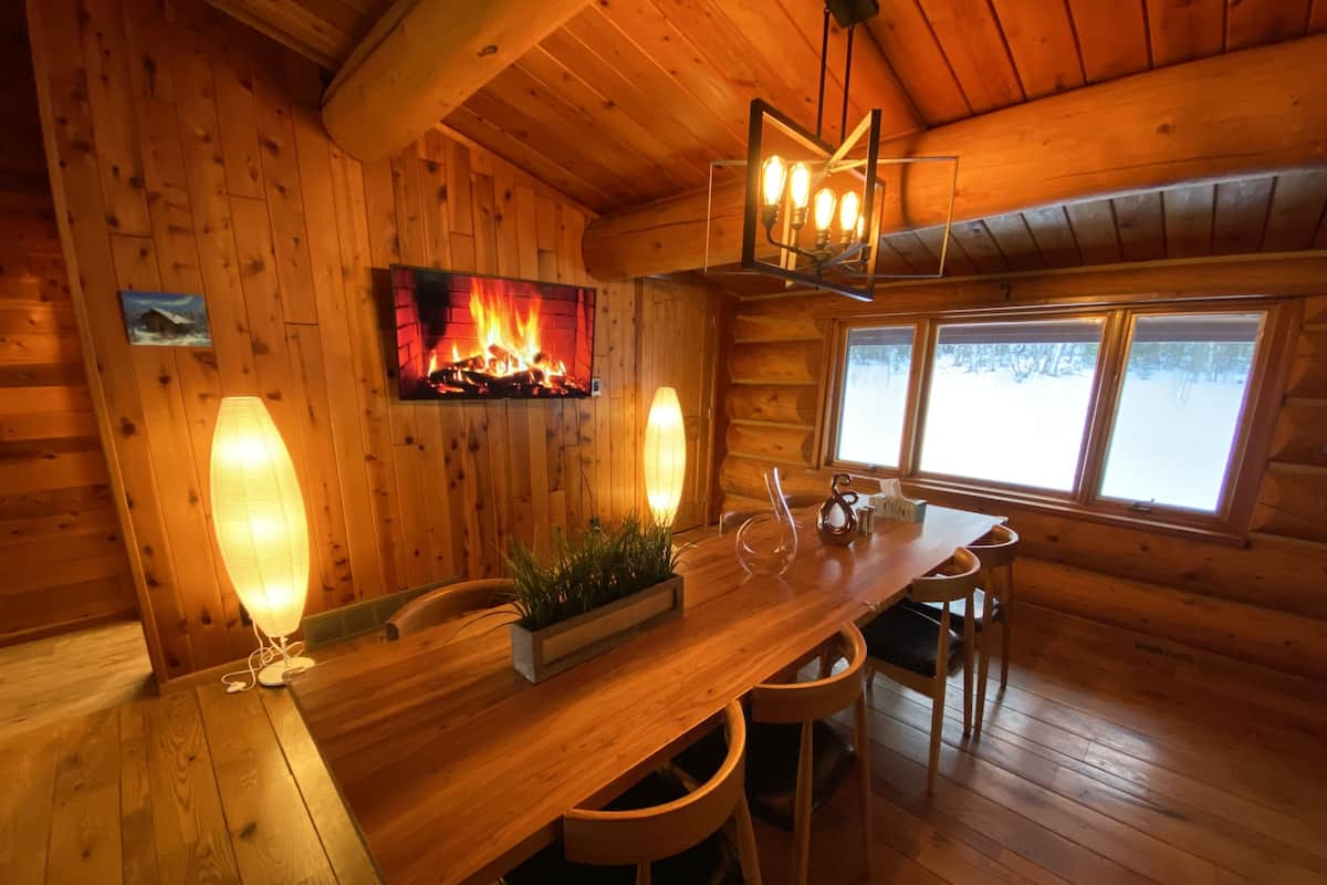 A cozy cabin interior with a wooden dining table, chairs, a fireplace, and a window with a snowy view.