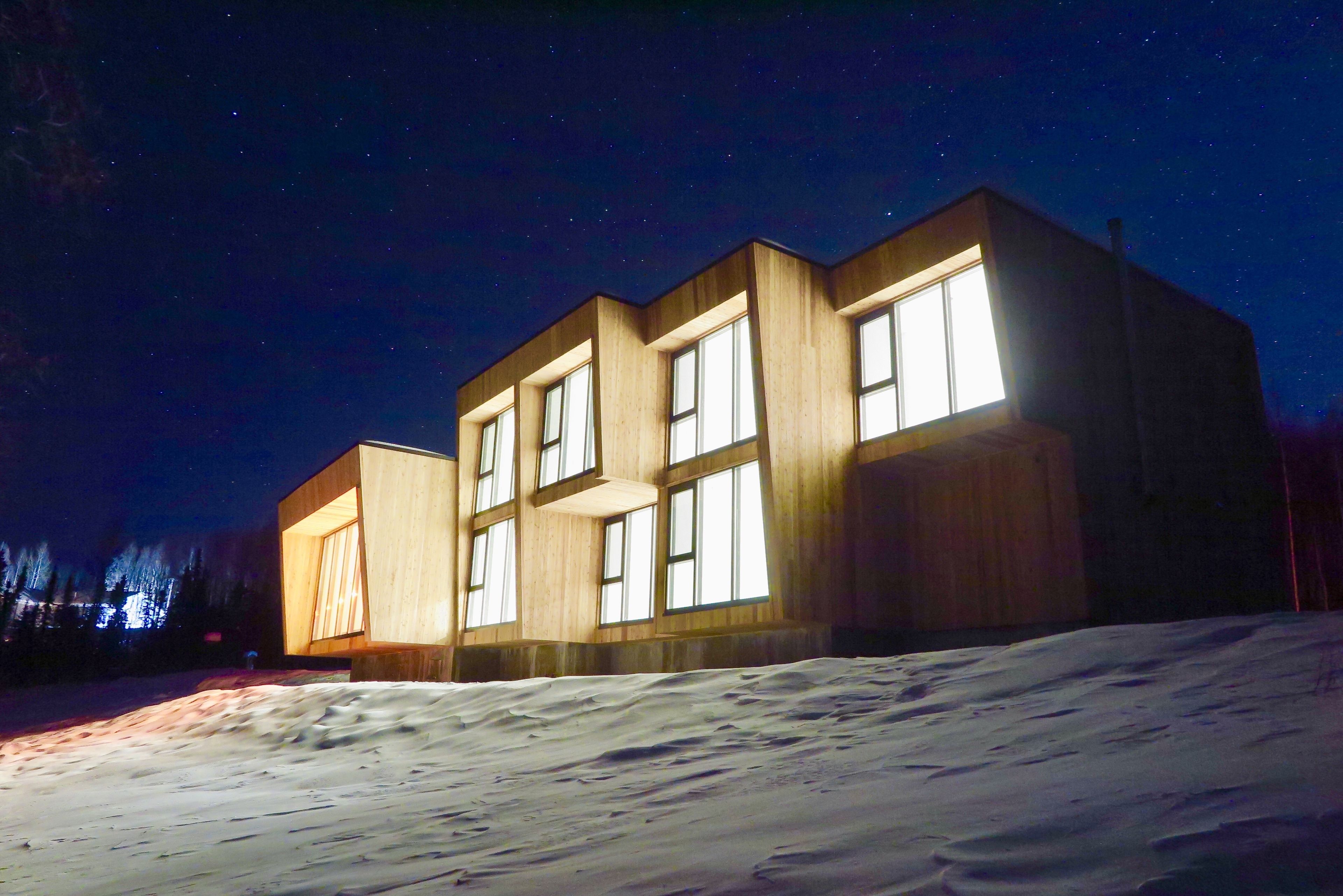 A modern wooden cabin with large windows, situated on a snowy slope under a starry night sky.