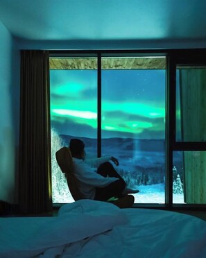 A room with a large window, a bed, and a person sitting by the window. - Aurora Villa (Fairbanks)