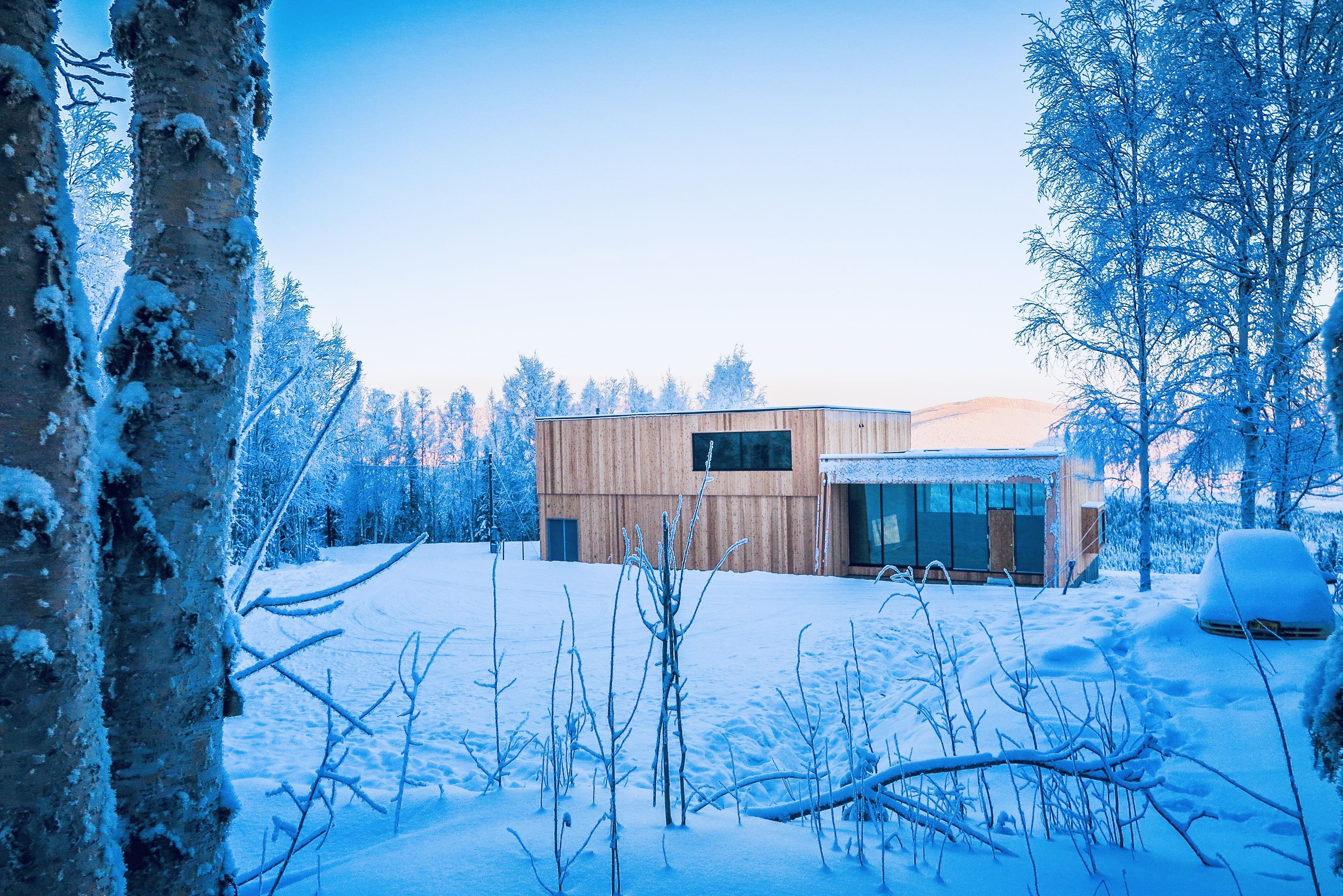 A modern wooden building with large windows and a glass door, surrounded by snow-covered trees.