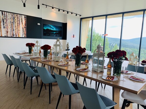 A dining area with a long table set for a meal, surrounded by chairs, with a large window offering a scenic view. - Aurora Villa (Fairbanks)