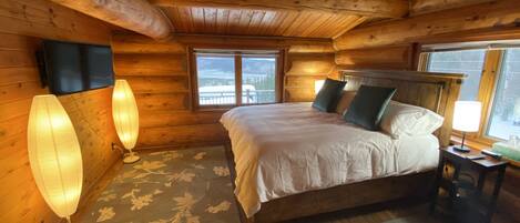 Quarto de cabana de madeira aconchegante, com uma cama grande, duas luminárias de cabeceira, uma cabeceira de madeira e vista para o exterior pela janela.