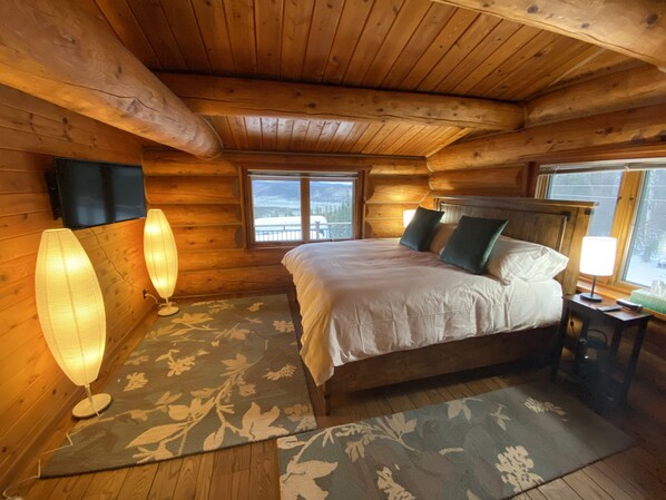 A cozy log cabin bedroom with a large bed, two bedside lamps, a wooden headboard, and a view of the outdoors through a window. - Aurora Villa (Fairbanks)