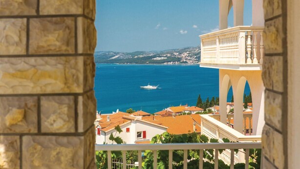 A coastal view from a balcony with a stone wall, a white building with arches, and a clear blue sea with a boat. - Gorgeous apartment in Okrug Gornji (Okrug Gornji)