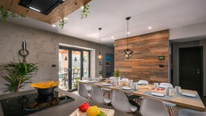 Un espace repas moderne avec une longue table, des chaises blanches et un îlot central doté d’une cuisinière et de légumes frais.