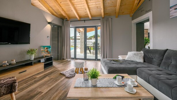 Un salon moderne avec un plafond en bois, un canapé gris, un téléviseur à écran plat et un coin repas avec une table mise pour deux personnes.
