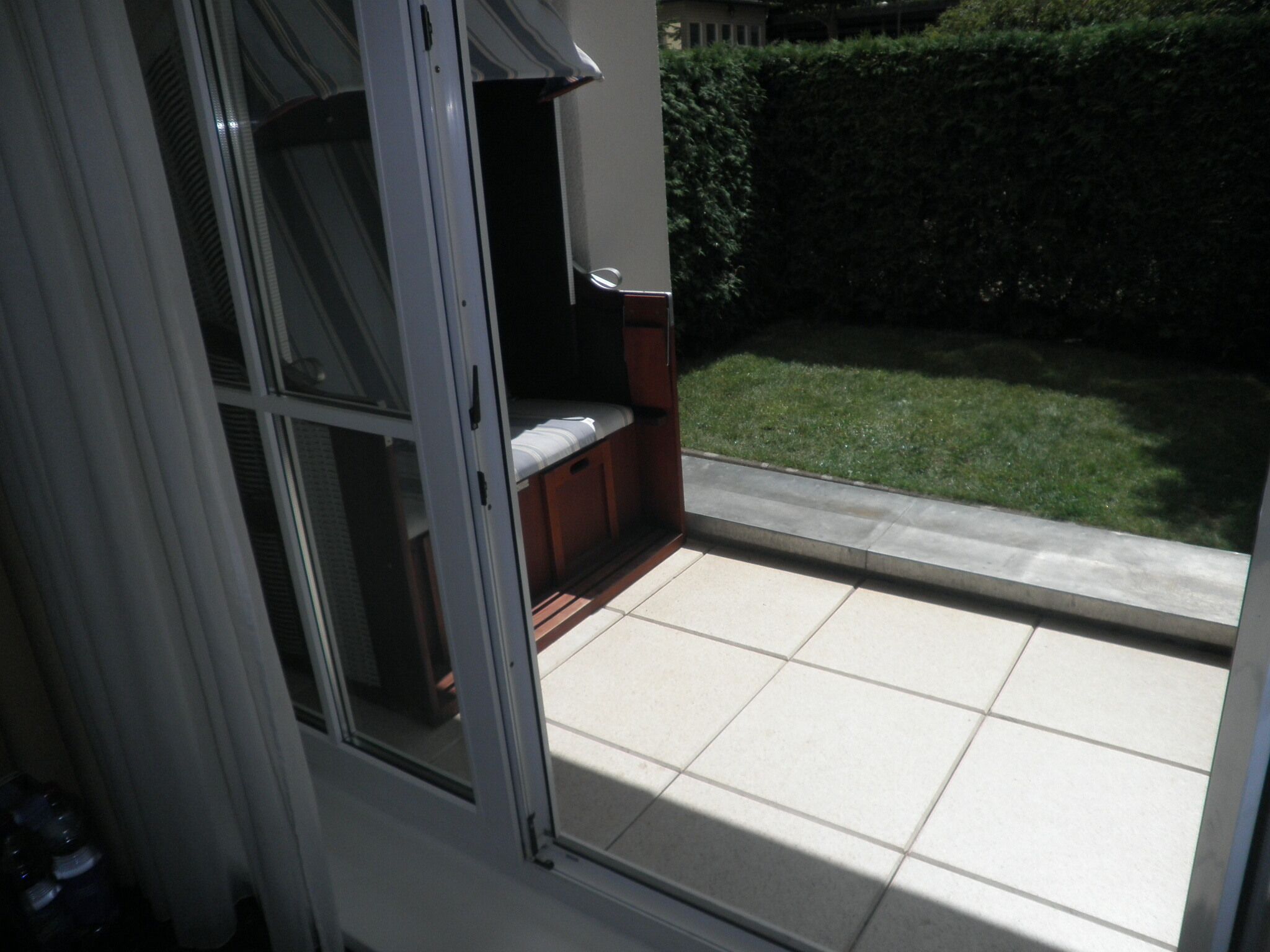 A view from a window with a tiled patio area, a wooden bench, and a hedge in the background.