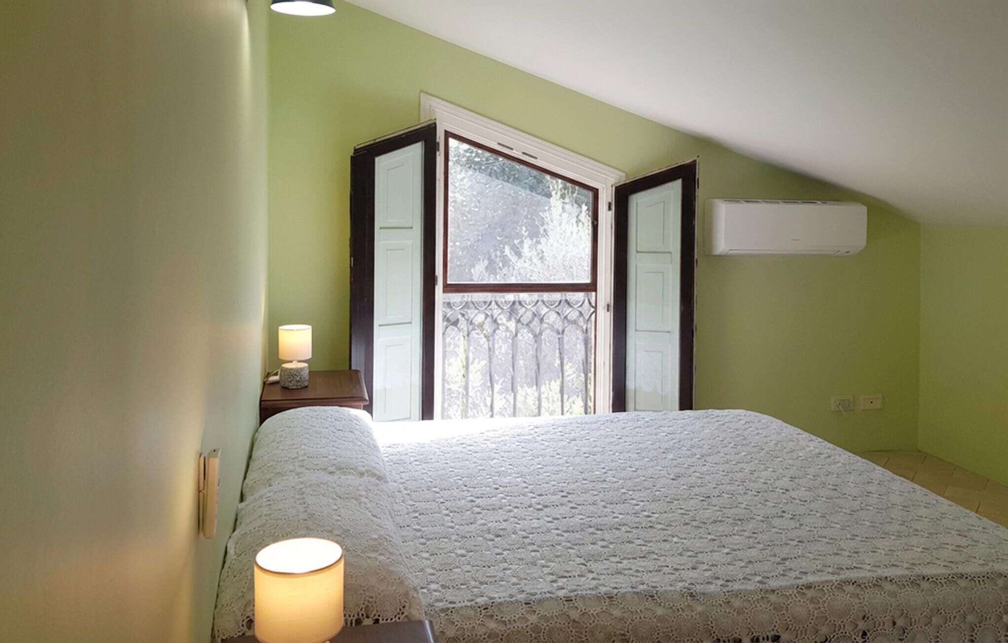 Un dormitorio con cama, mesita de noche con lámpara, ventana con vista a árboles y un equipo de aire acondicionado en la pared.
