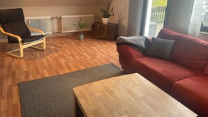A modern living room with a wooden floor, a red sofa, a black chair, a wooden coffee table, a radiator, a potted plant, and a sliding glass door leading to an outdoor area. - Very nice apartment with a large sun terrace (Stadtkyll)