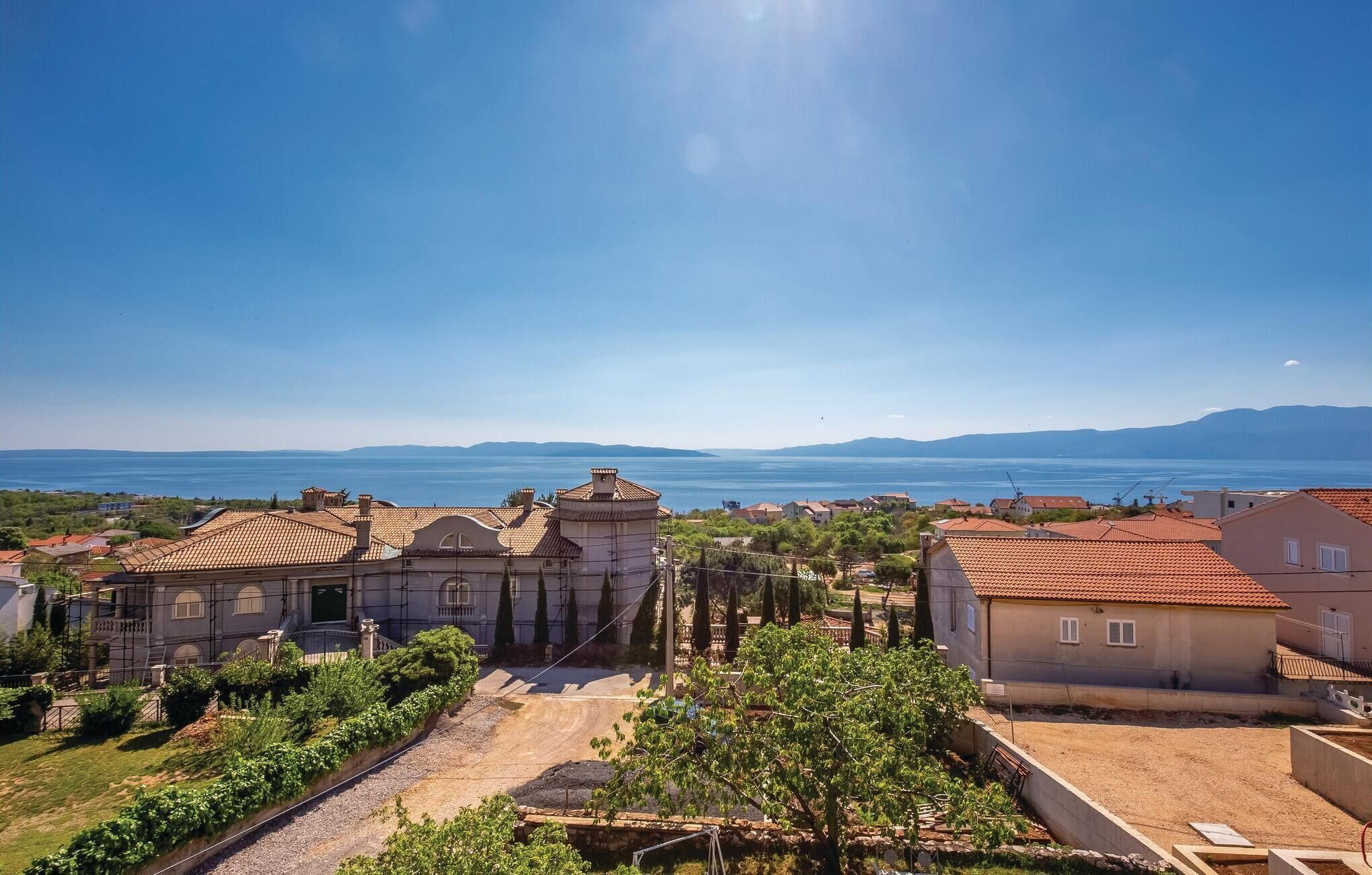A coastal town with terracotta-roofed buildings, a clear blue sky, and a view of the sea and distant mountains.