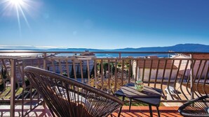 Un balcón con sillas de mimbre y una mesa, con vistas a la costa, edificios y montañas a lo lejos.