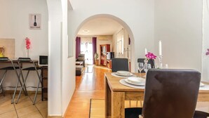 Un comedor con una mesa de madera dispuesta para cuatro, un televisor al fondo y una zona de cocina con un adorno de espátula en la pared.