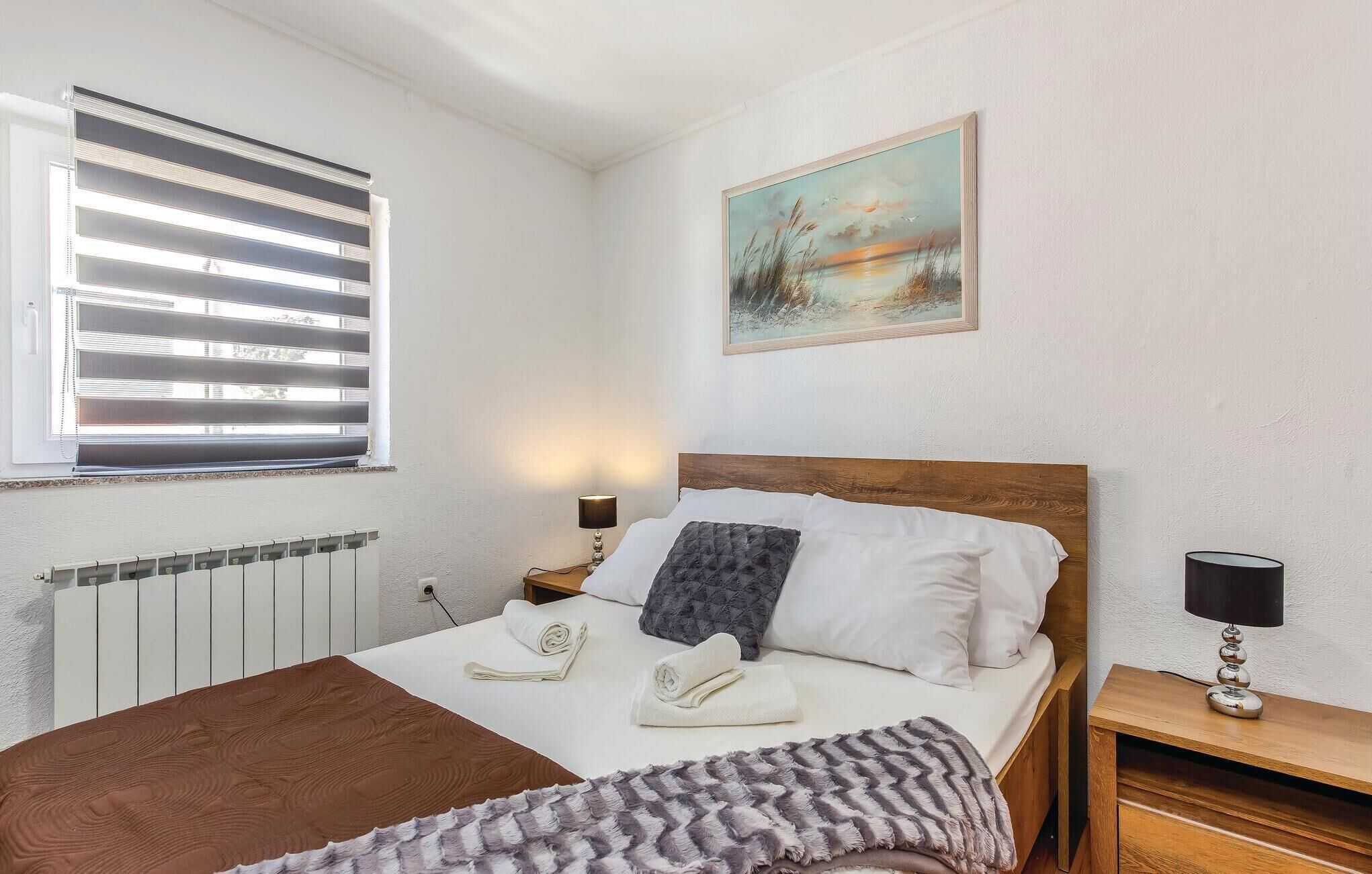 Ein Schlafzimmer mit einem hölzernen Bett, weißer Bettwäsche, einem Fenster mit Jalousien, einem Heizkörper, einem Bild an der Wand und einem Nachttisch mit einer Lampe.