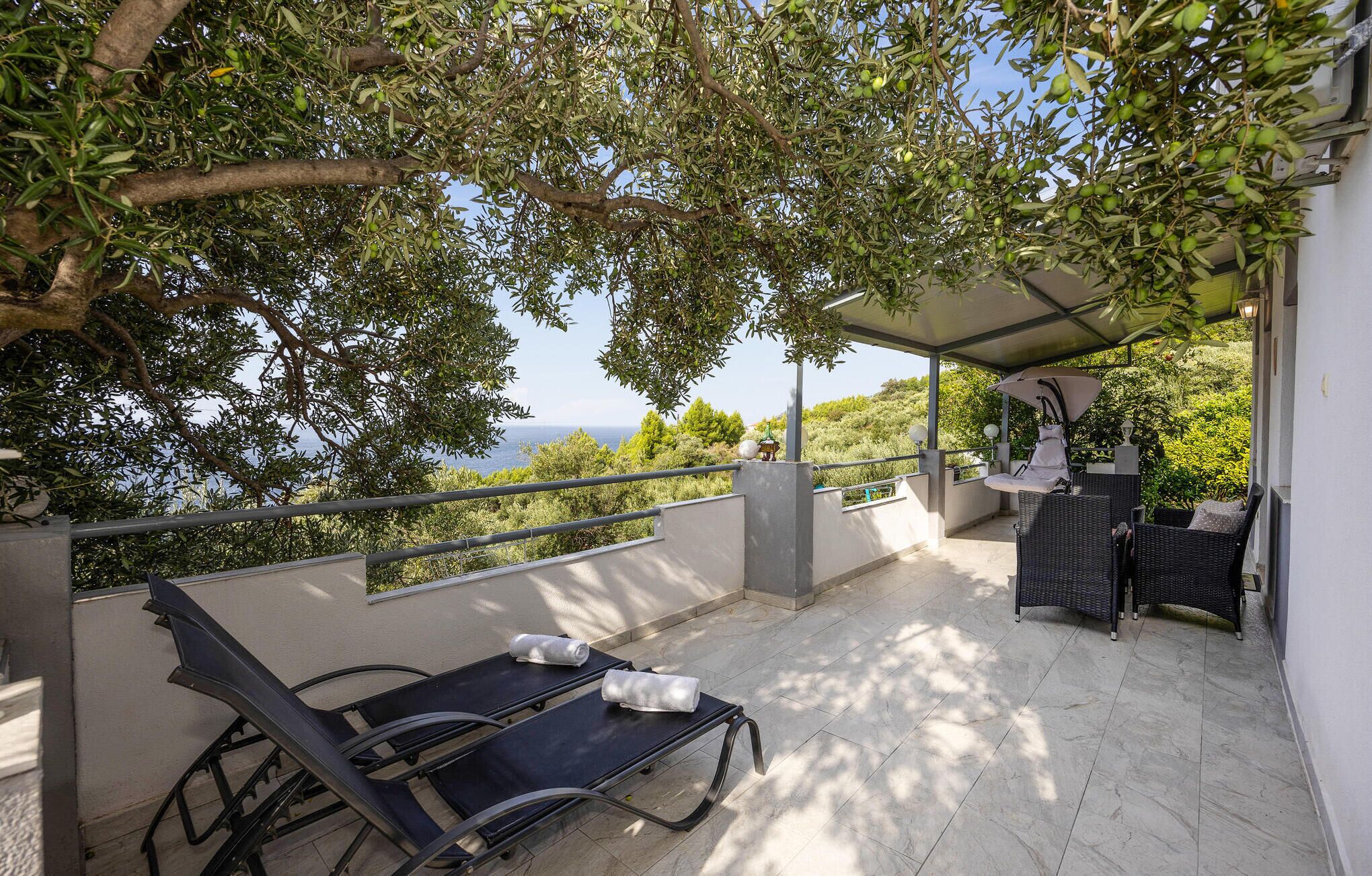 Eine Terrasse mit zwei Liegestühlen, einem Tisch und einem Schaukelstuhl, mit Blick auf eine schöne Aussicht mit Bäumen und dem Meer.