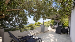 A terrace with two lounge chairs, a table, and a swing chair, overlooking a scenic view with trees and the sea.
