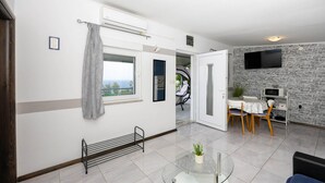 A modern living room with a glass coffee table, a dining area with a table and chairs, a TV mounted on a stone wall, and a view through a window.