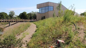 A modern building with a unique wooden exterior and large windows, situated on a hillside with a dirt path leading to a wooden bridge. - Barrier-free relaxation in a quiet location near the Elbe, the Altmarkt (Sandau/Elbe)