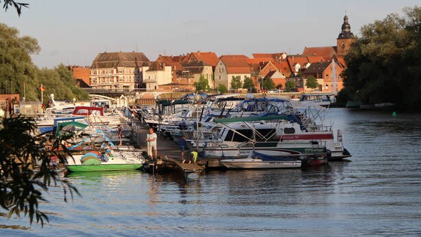 A marina with boats docked, a bridge in the background, and buildings with red roofs. - Barrier-free relaxation in a quiet location near the Elbe, the Altmarkt (Sandau/Elbe)