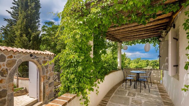 Une terrasse aux murs de pierre, dotée d’un sol carrelé, d’une pergola recouverte de plantes grimpantes et d’une table avec des chaises.