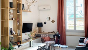 A modern living room with a dining area, wooden shelving, a flat-screen TV, and a large window with curtains. - Home Chic Home - Les Suites de La Comédie - Suite2 (Montpellier)