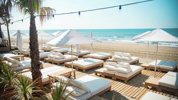 A beachside lounge area with wooden loungers, white cushions, and umbrellas providing shade.