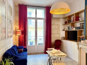 Een woonkamer met een blauwe bank, een tafel met boeken, een rode fauteuil, een boekenkast en een groot raam met gordijnen.