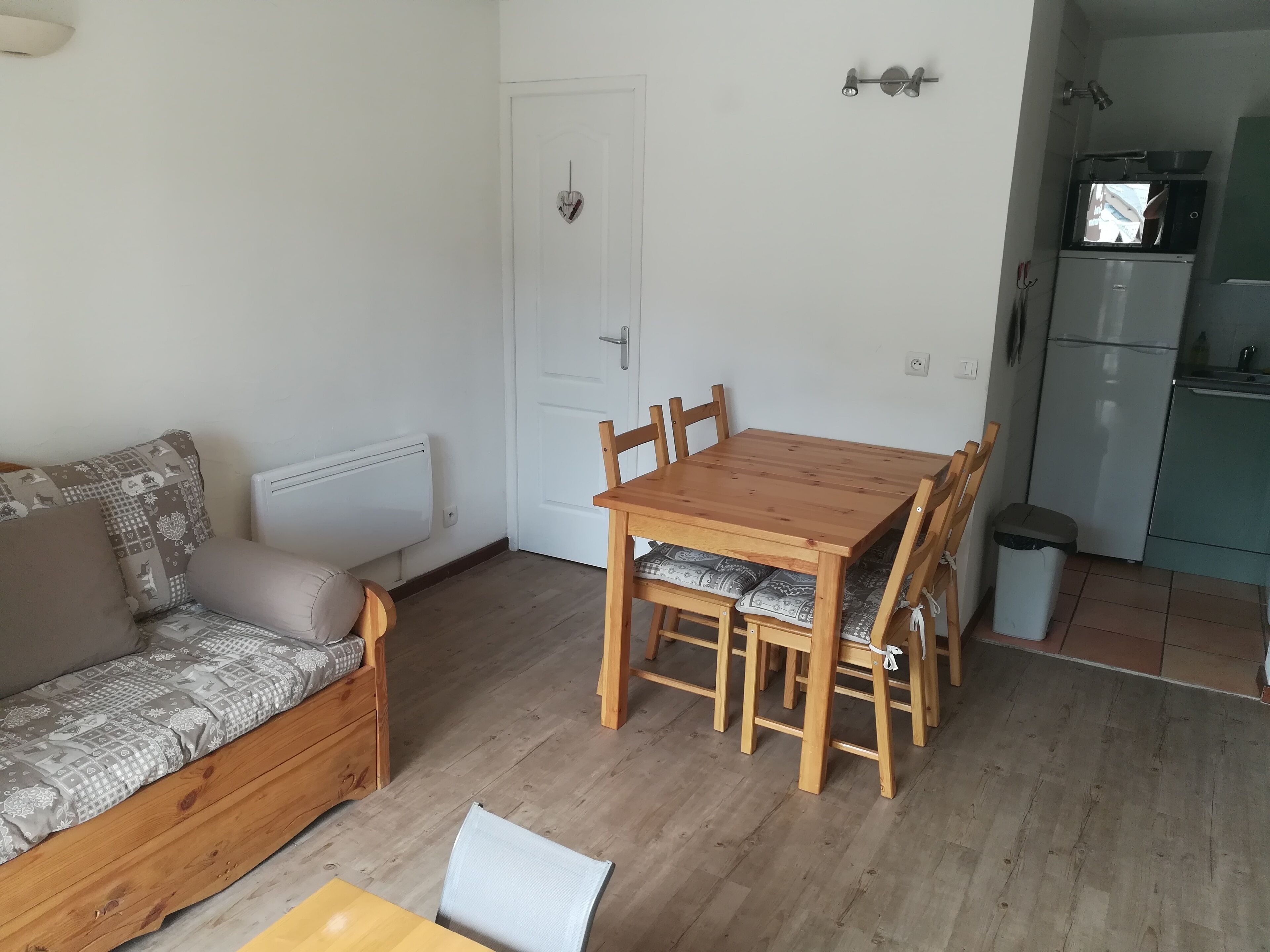 A compact living space with a wooden dining table and chairs, a sofa with patterned cushions, a white door, and a kitchenette with a refrigerator and microwave.