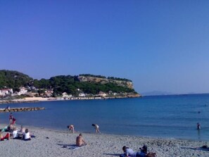 A beach with people sunbathing and swimming, a rocky cliff with buildings, and a clear blue sky.