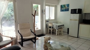 A living room with a glass coffee table, a sofa, a chair, and a dining area with a table and chairs. There is a refrigerator and a television in the kitchen area. - Garden level apartment, for 5 persons, beach et lake 5 min away, great amenities (Soorts-Hossegor)