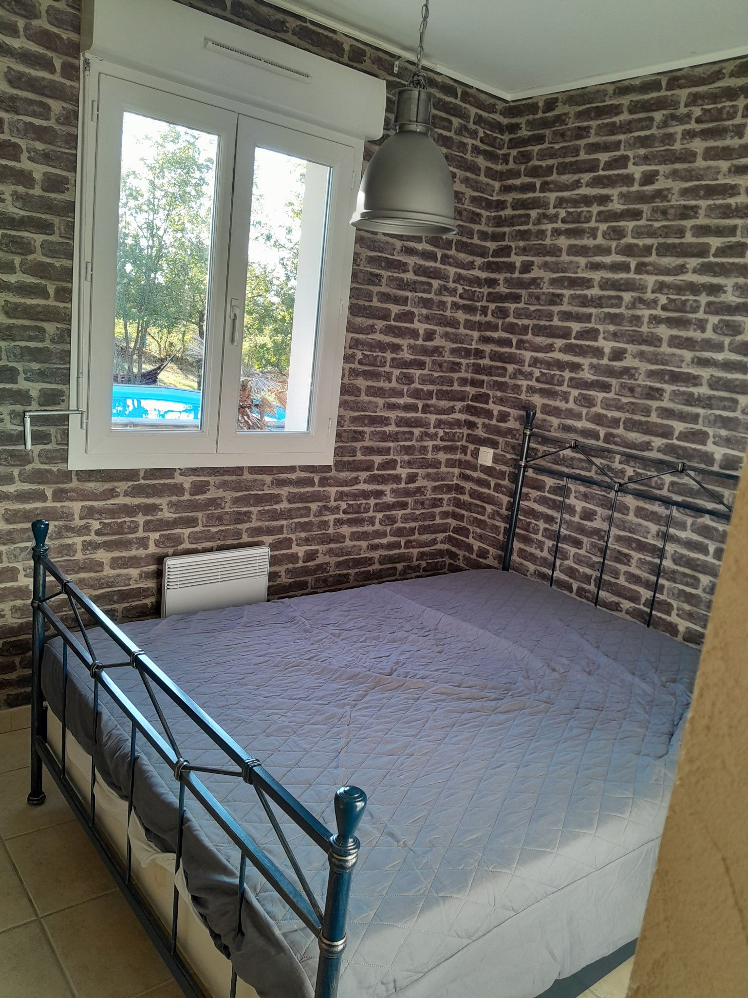 Una habitación con pared de ladrillo, cama con estructura de metal, ventana con vistas a vegetación y una lámpara colgante.