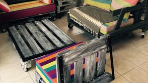 Une pièce comprenant un banc en bois, un fauteuil rembourré et une petite table. On y trouve une fenêtre avec des stores, une plante en pot et une lampe fixée au mur.